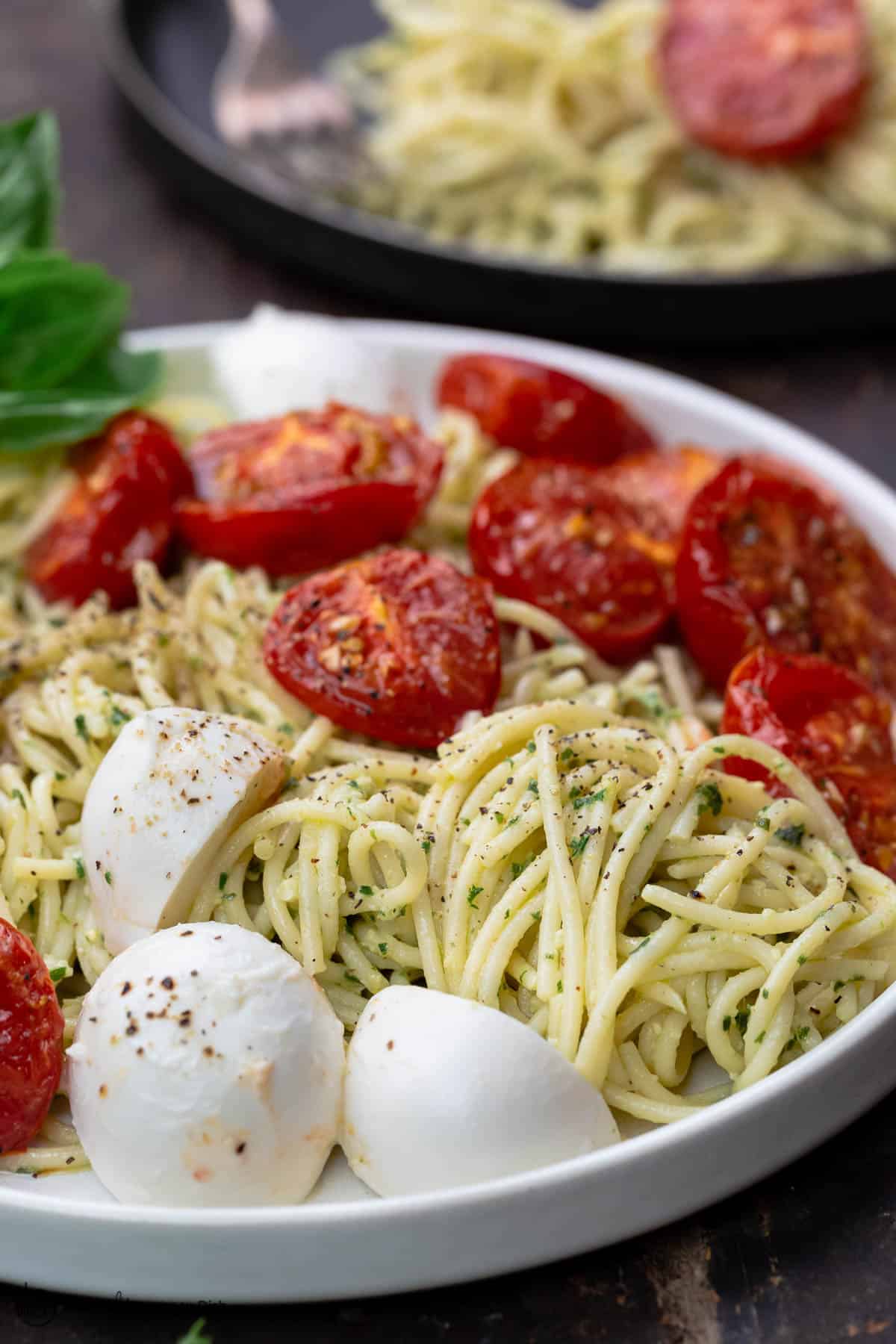 Pasta in a large bowl, tossed with pesto, tomatoes and mozzarella. A small plate to the side