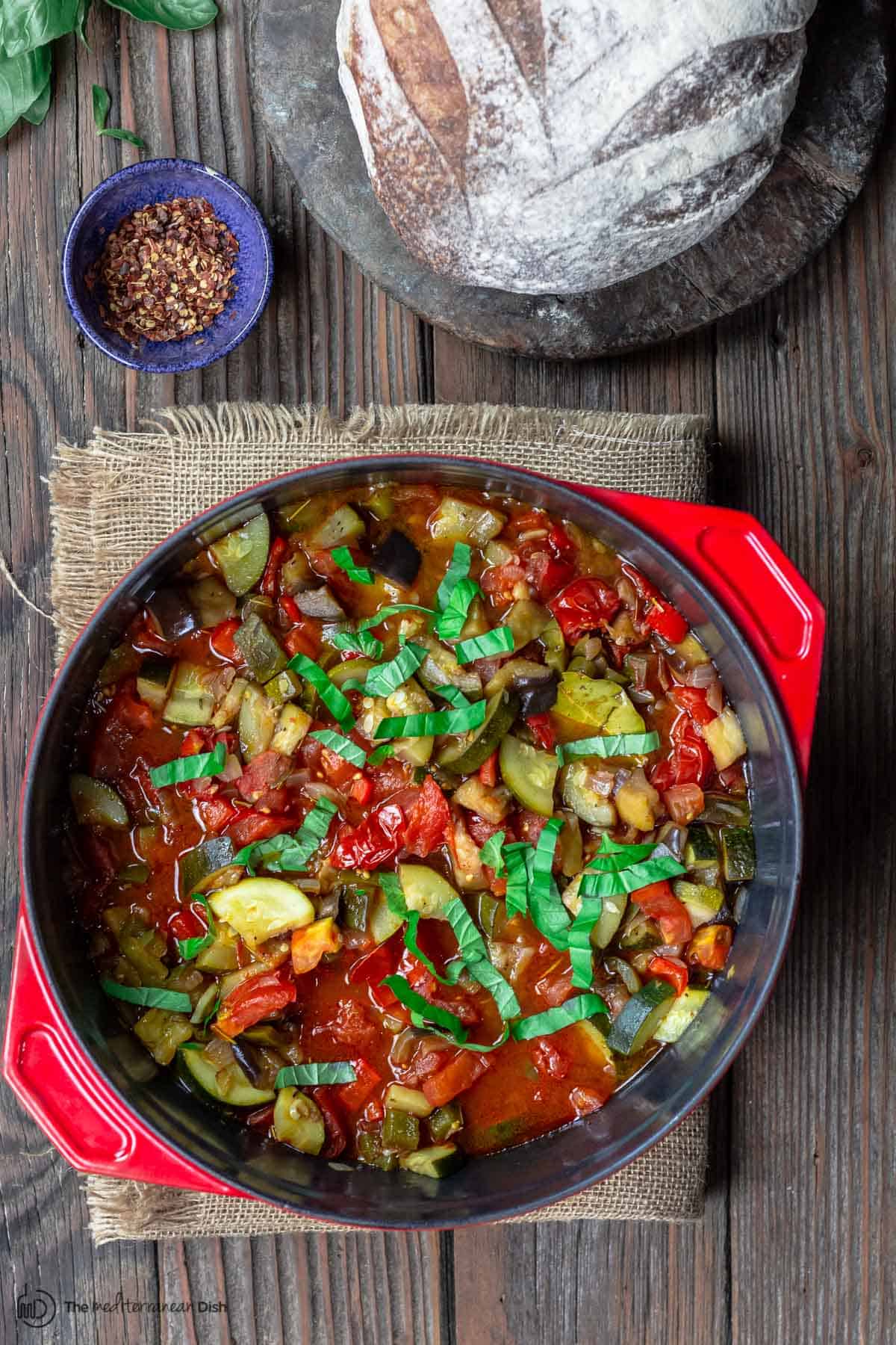 Rataouille served in a large red pot on a table with a loaf of bread