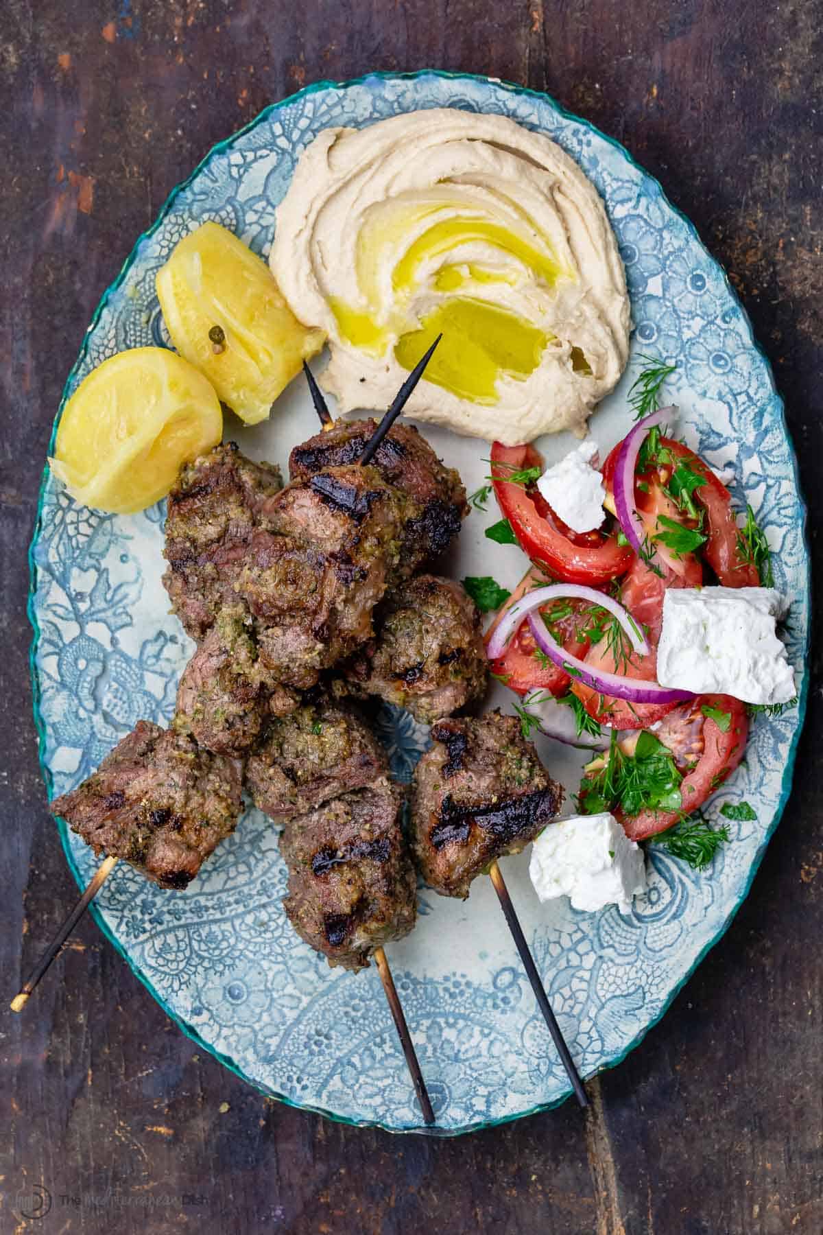 Lamb kabobs served on a plate with veggies and hummus