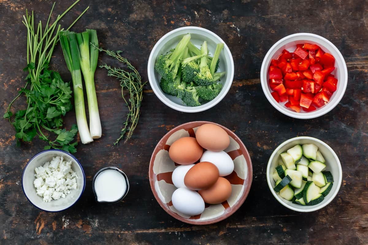 ingredients. Seven Eggs, A Bowl of Diced Red Bell Pepper and the Remaining Frittata Ingredients on a Dark Wooden Surface