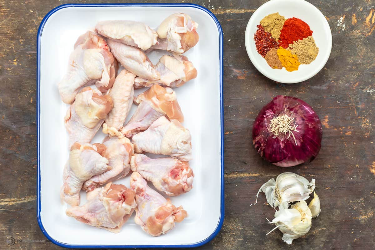 Raw chicken wings on a tray with a dish of spices, an onion and garlic cloves