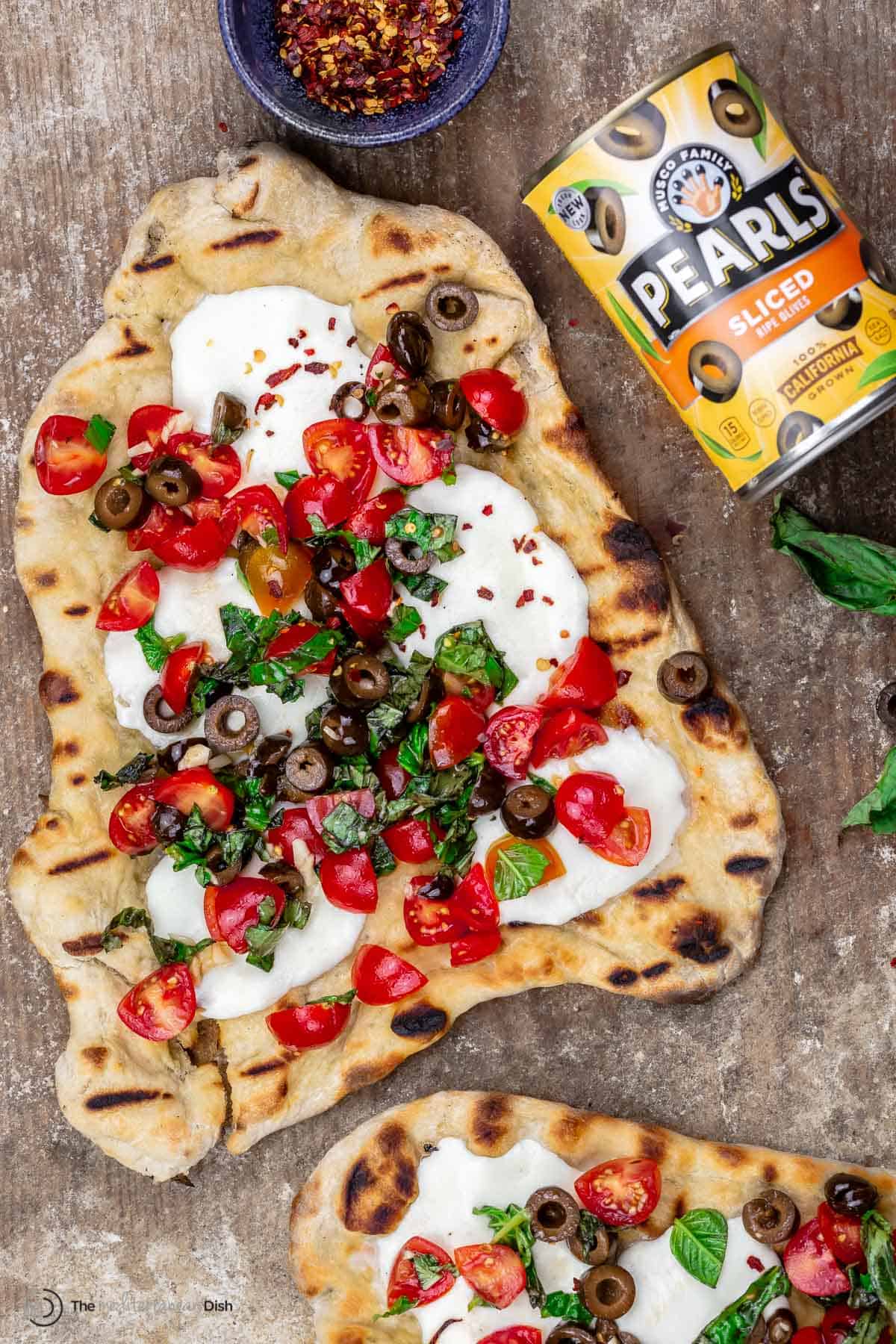 grilled pizza on wood board with a side of red pepper flakes and a can of Pearls Olives