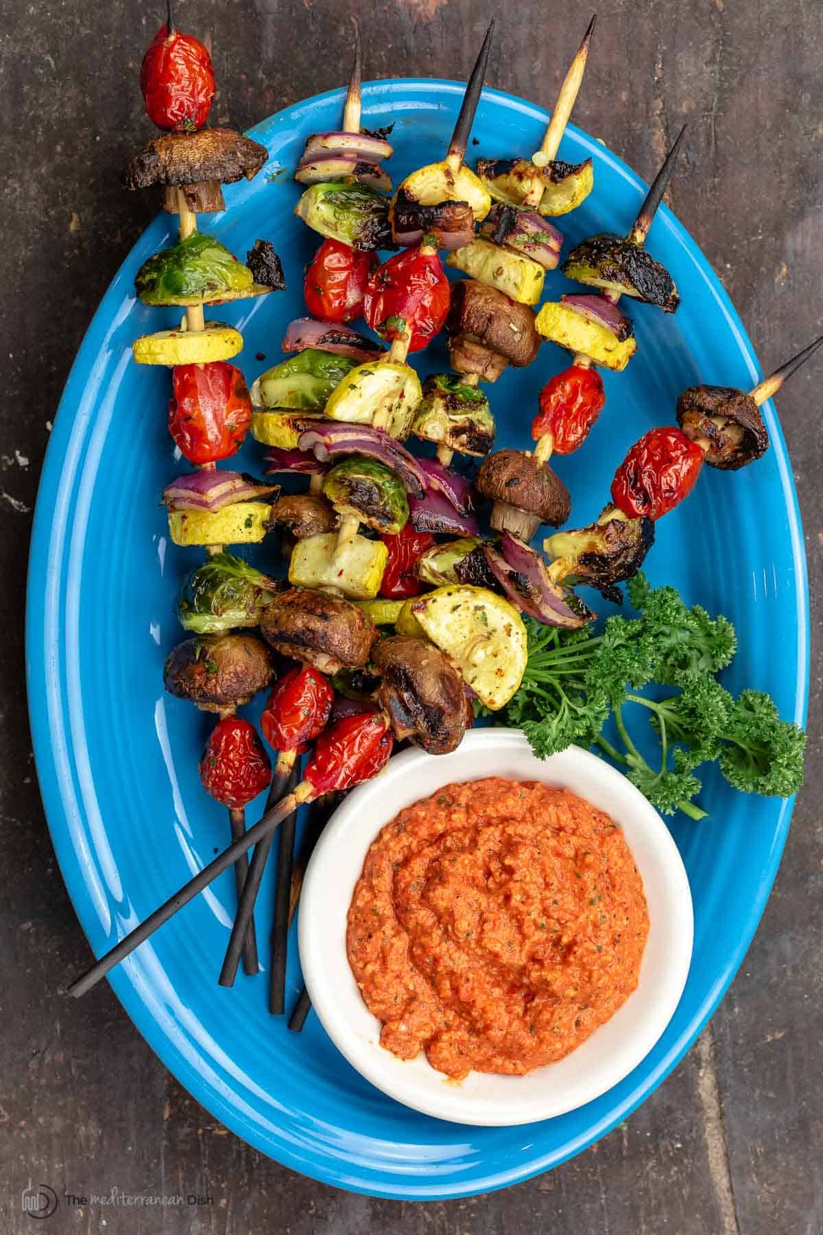 Grilled veggie kabobs on a blue plate with a dish of romesco sauce