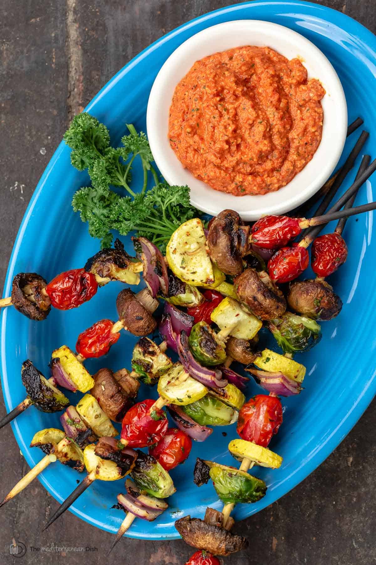 Overheard view of grilled vegetable skewers on a blue plate with a dish of romesco sauce