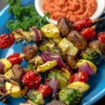 A blue plate with several grilled vegetable kabobs and a dish of romesco sauce