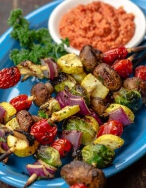 A blue plate with several grilled vegetable kabobs and a dish of romesco sauce