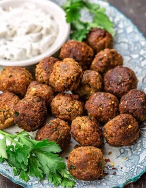 Eggplant meatballs on a blue plate, with a dish of tzatziki and garnished with parsley