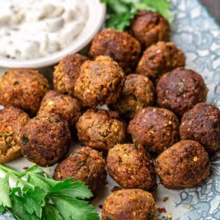 Eggplant meatballs on a blue plate, with a dish of tzatziki and garnished with parsley