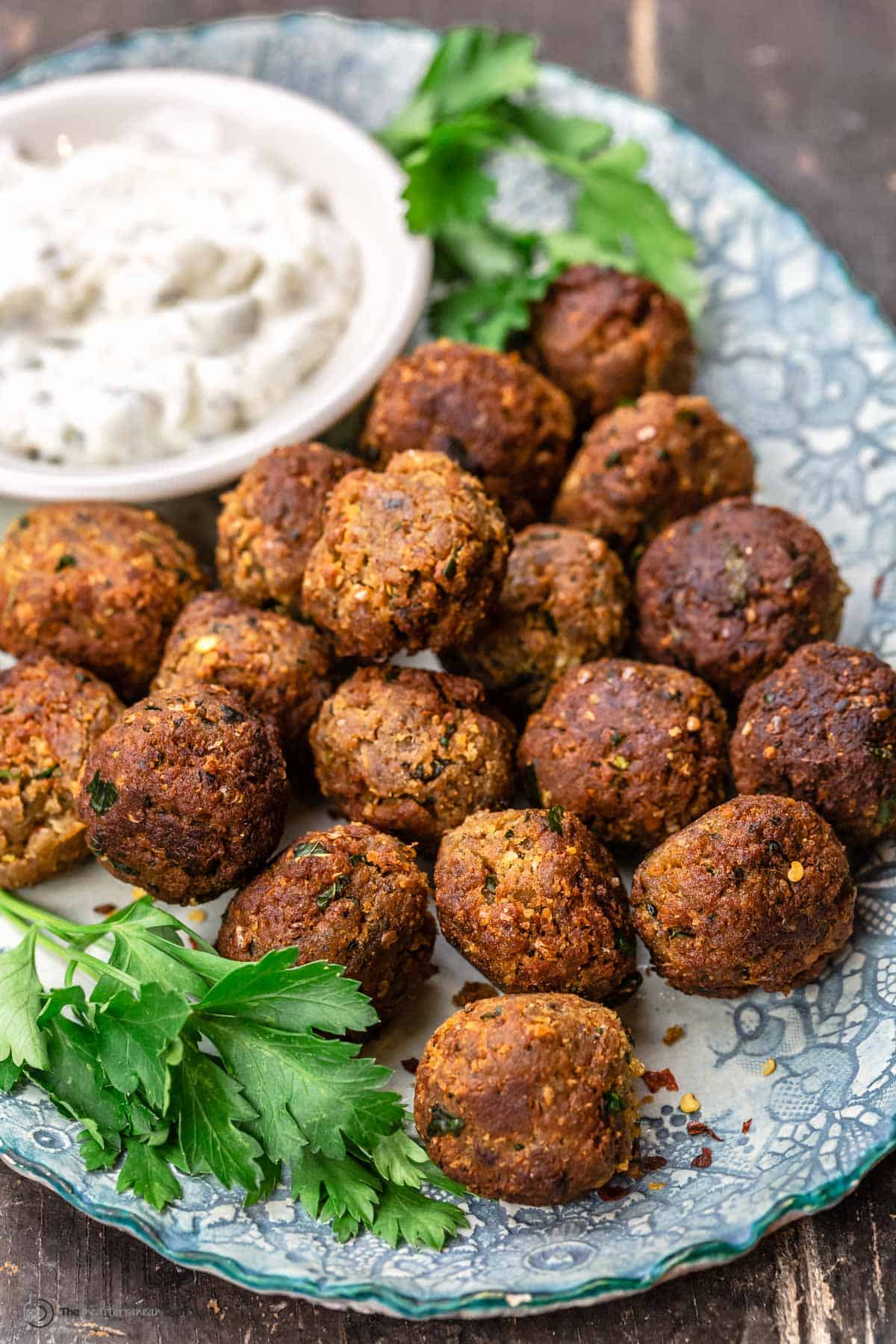 Eggplant meatballs on a blue plate, with a dish of tzatziki and garnished with parsley