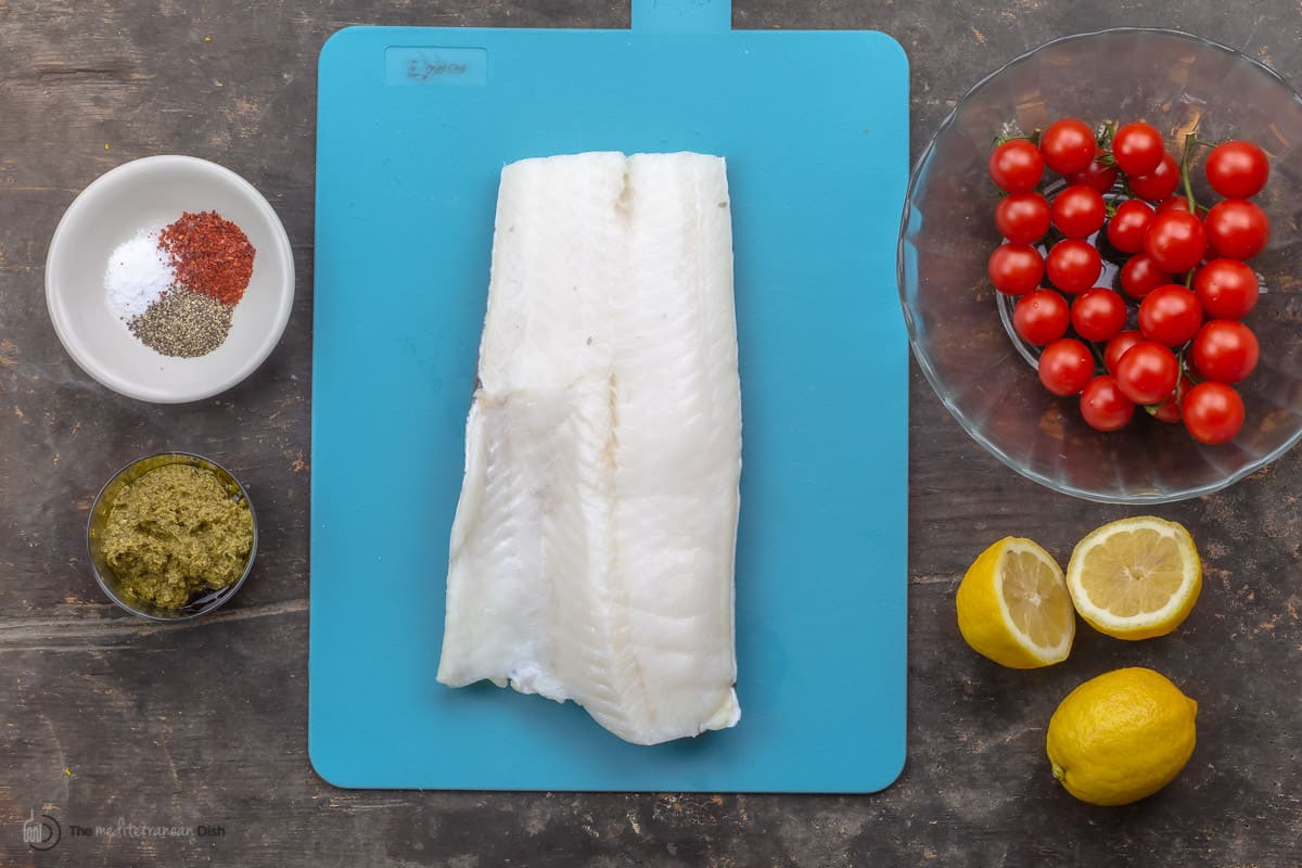 A sea bass fillet on a cutting board, a bowl of grape tomatoes, lemon halves and seasonings