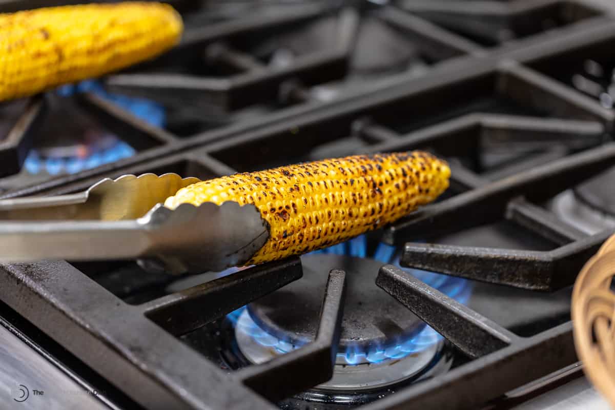 An ear of corn being turned over the flame of a gas burner