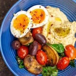 A Mediterranean breakfast bowl with a soft-boiled egg, hummus and sauteed vegetables on a blue plate.