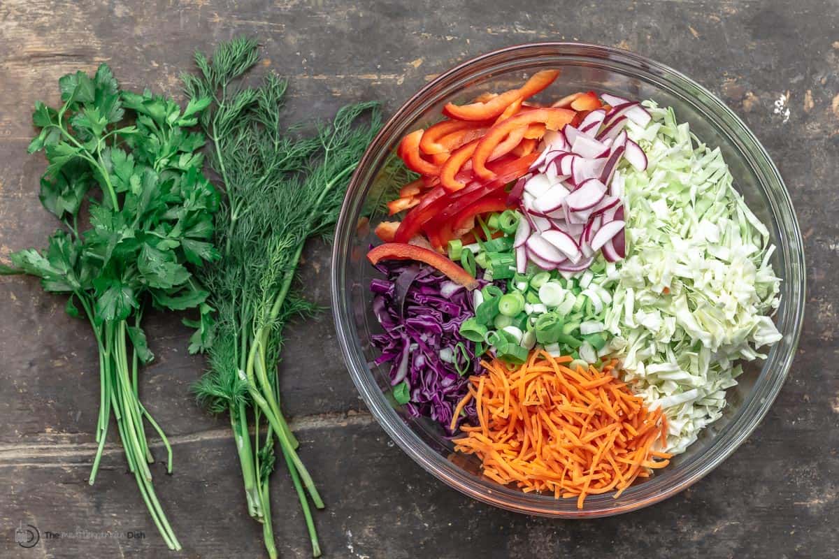 A bowl of chopped veggies - red peppers, cabbage, carrots, radishes - next to two bunches of fresh herbs