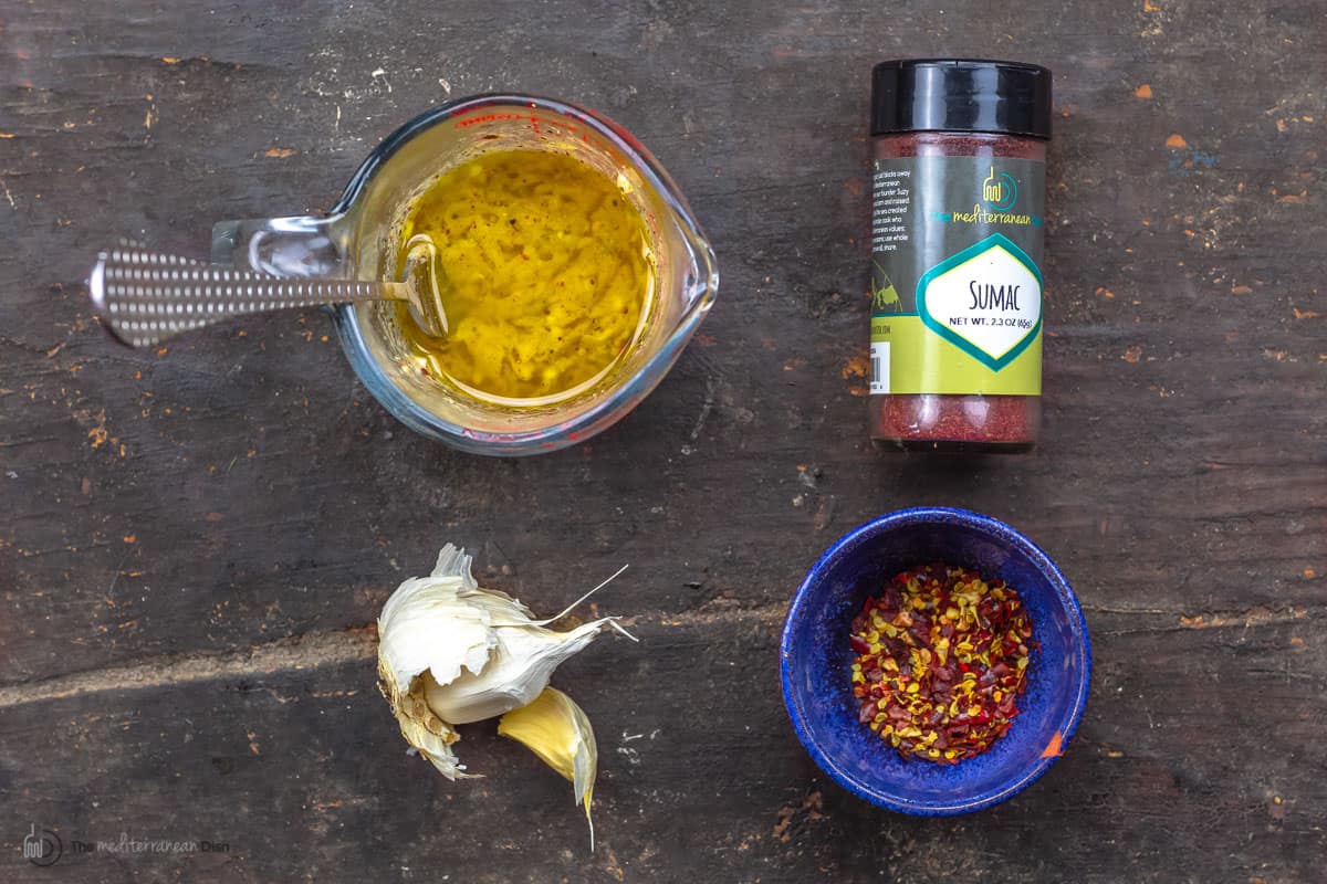 A measuring cup with dijon mustard and lemon juice, a shaker of Sumac, garlic and a dish of spices