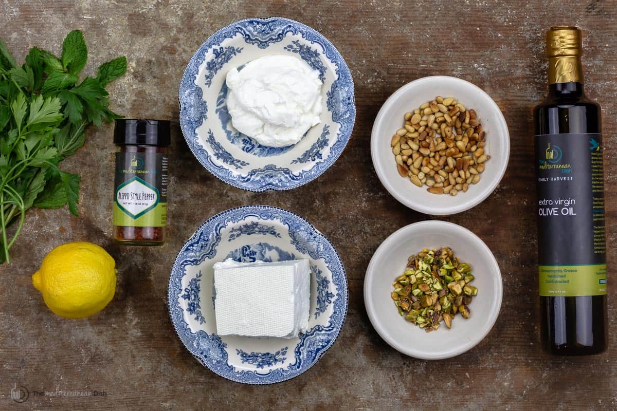 Greek yogurt, feta cheese, pine nuts and crushed pistachios measured into bowls with a lemon, aleppo pepper and olive oil on the sides