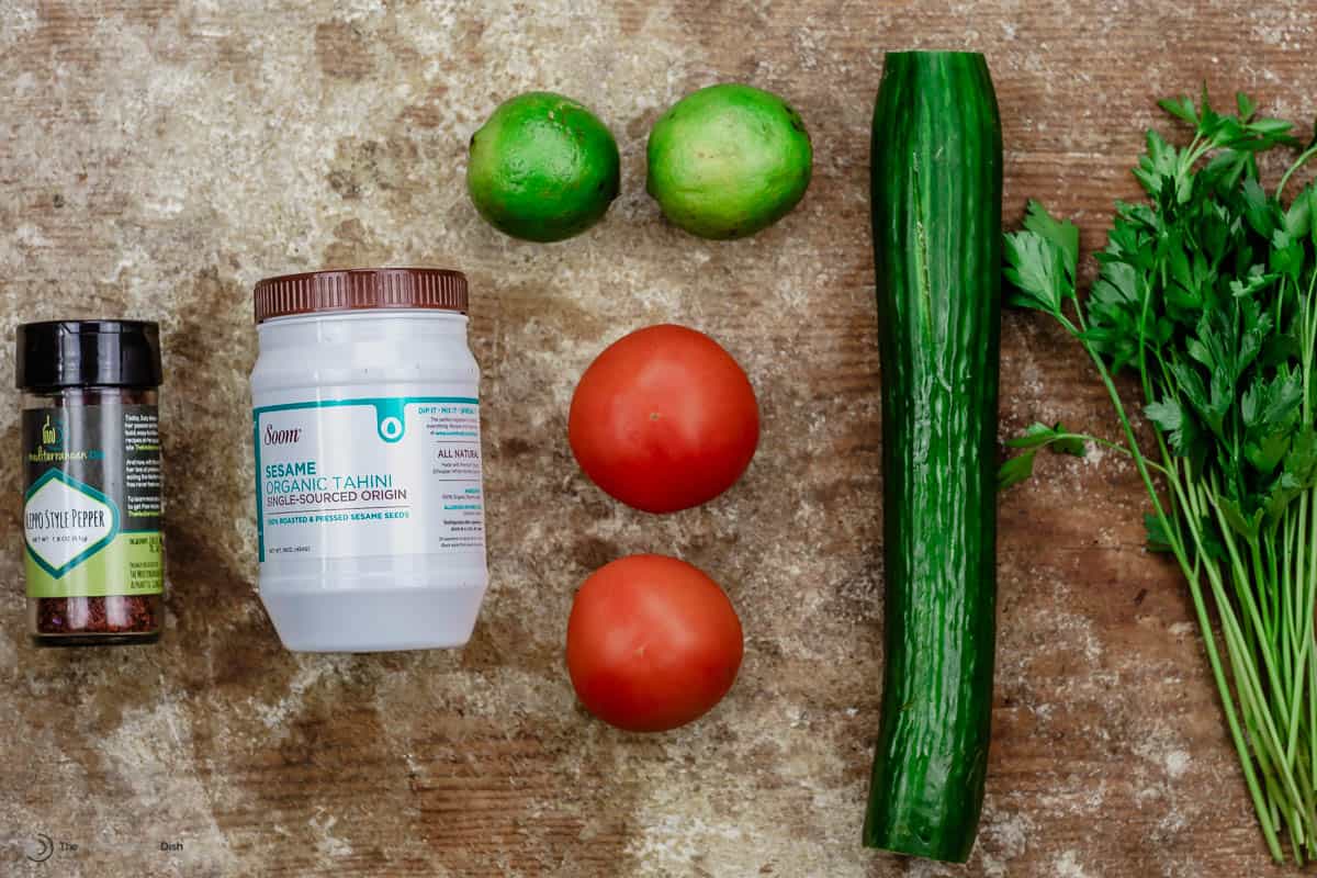 A jar of Aleppo pepper, tahini, two limes, two tomatoes, a cucumber and fresh parsley