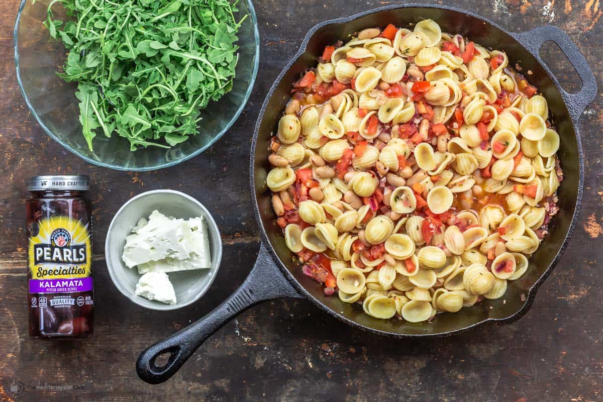 cooked pasta with sauce in the skillet with a bowl of arugula and a bowl of feta to the side and a jar of kalamata olives