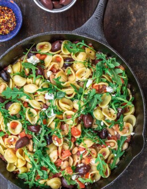 Orecchiette pasta in a skillet, topped with arugula. smaller bowls of red pepper flakes and olives to the side