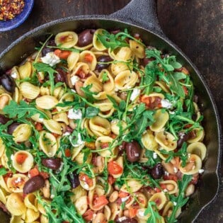 Orecchiette pasta in a skillet, topped with arugula. smaller bowls of red pepper flakes and olives to the side