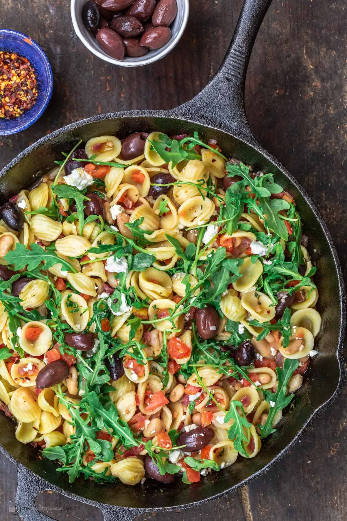 Orecchiette pasta in a skillet, topped with arugula. smaller bowls of red pepper flakes and olives to the side