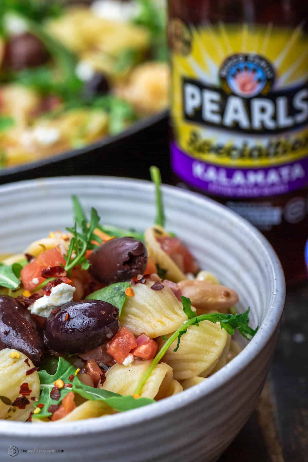 bowl of white bean orechiette pasta with a jar of olives in the background