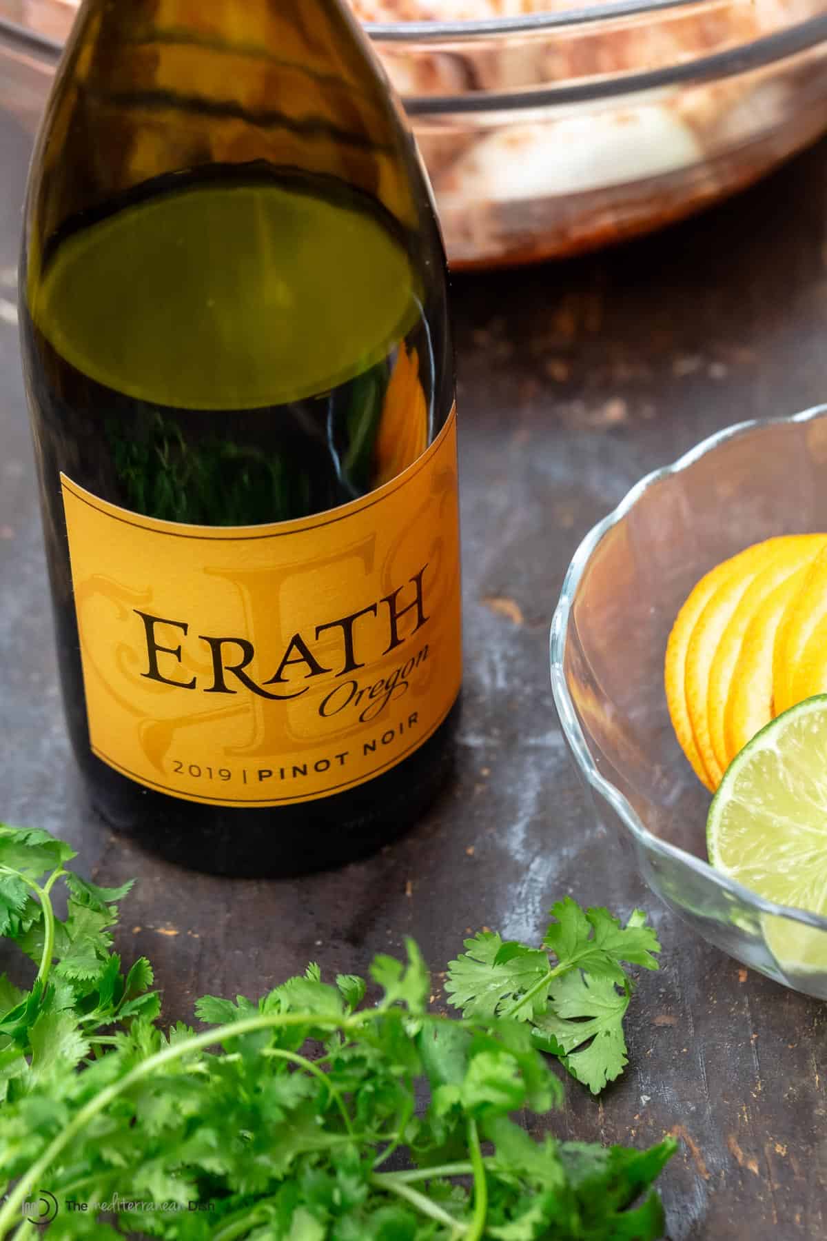 Marinated chicken in a bowl, and a side bowl with sliced citrus fruit, a bottle of wine and some fresh parsley on the side