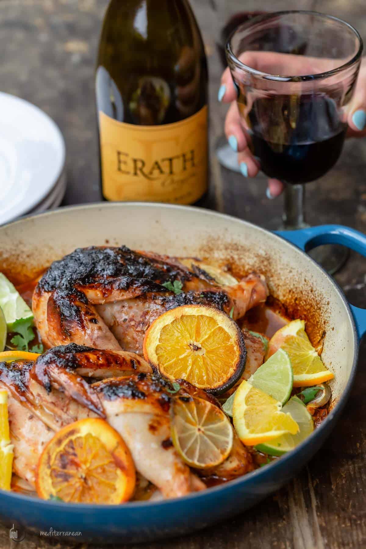 Roasted chicken with rosemary and citrus in a braising pan with a bottle of wine and a glass of wine held by a hand next to it