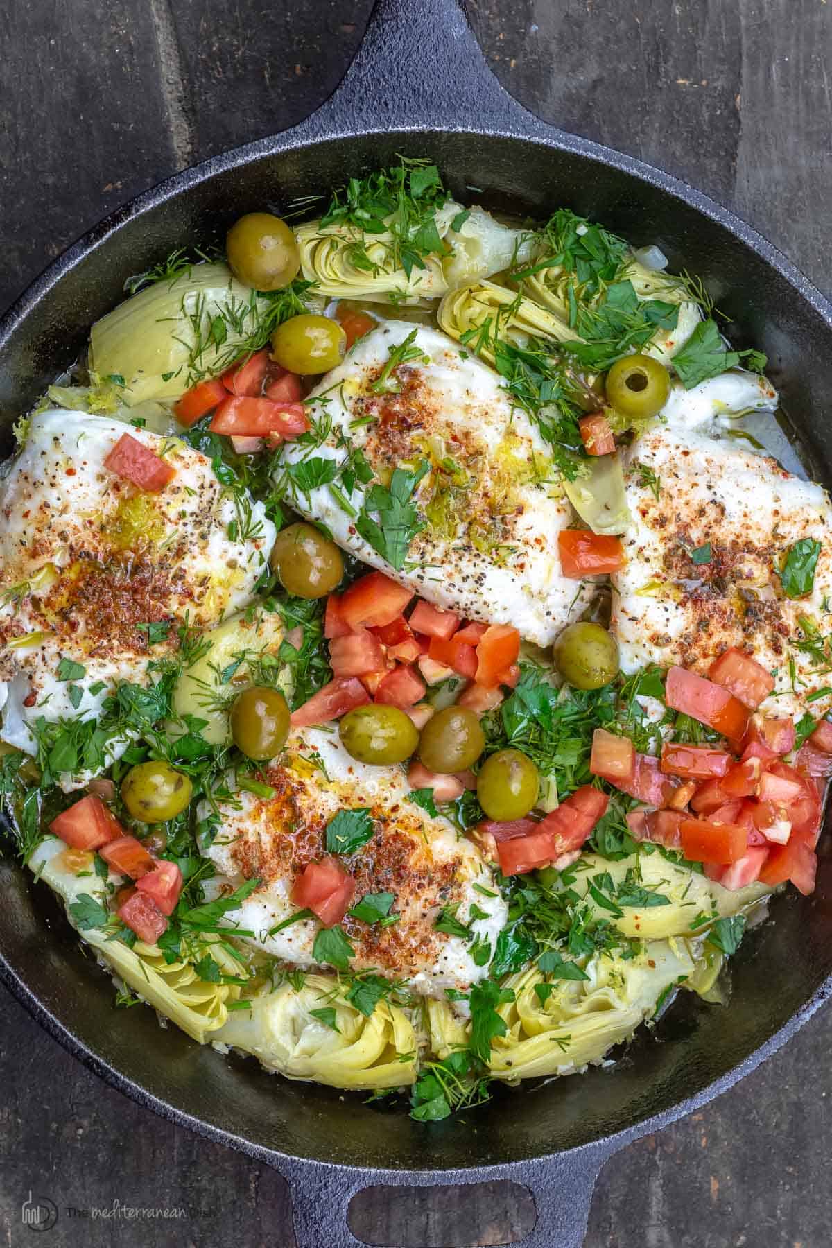 Overhead view of lemon haddock fillets with artichoke hearts and tomatoes in a skillet
