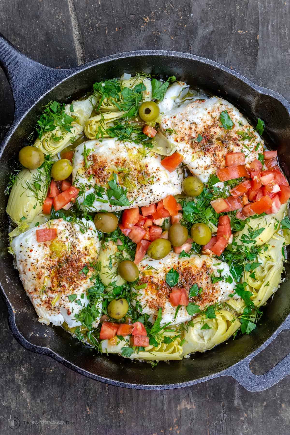 Overhead view of lemon haddock fillets with artichoke hearts and tomatoes in a skillet