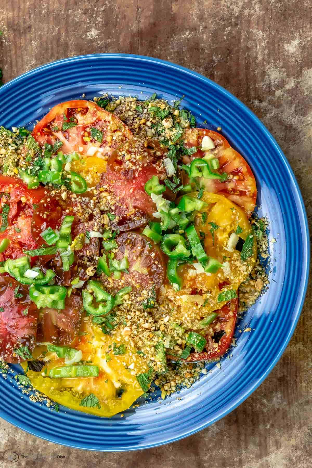 Heirloom tomato salad on a blue plate, topped with dukkah and jalapeno peppers