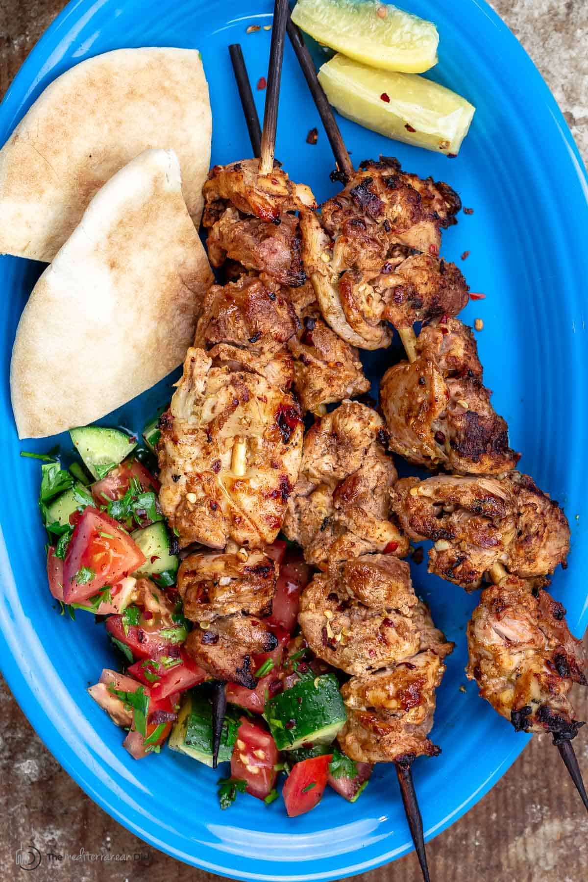 grilled shish tawook skewers on a large blue platter with a side salad, preserved lemons, and pita wedges