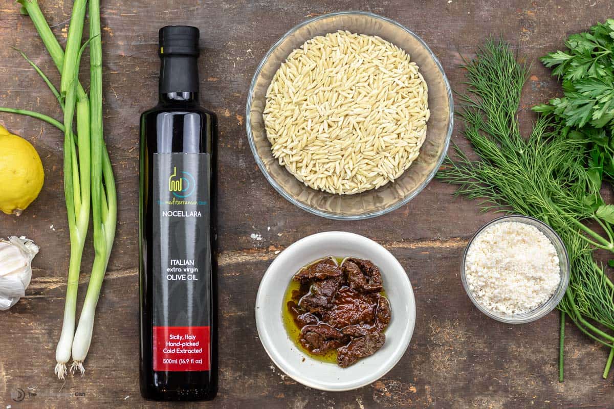 Overhead view of ingredients for orzo recipe including orzo, sundried tomatoes, parsley, parmesan cheese, green onions, garlic and lemon