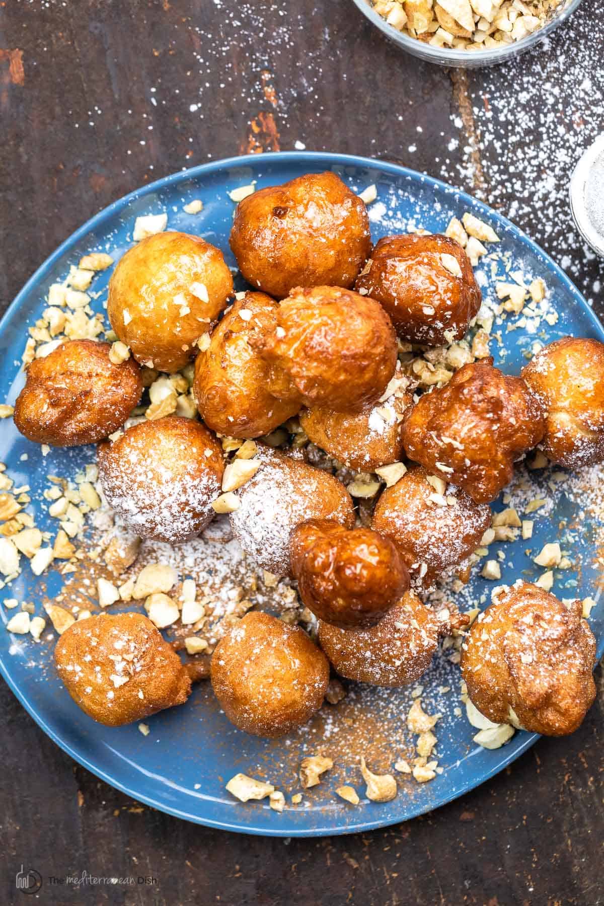 A blue plate full of loukoumades topped with confectioners' sugar, crushed nuts and honey syrup