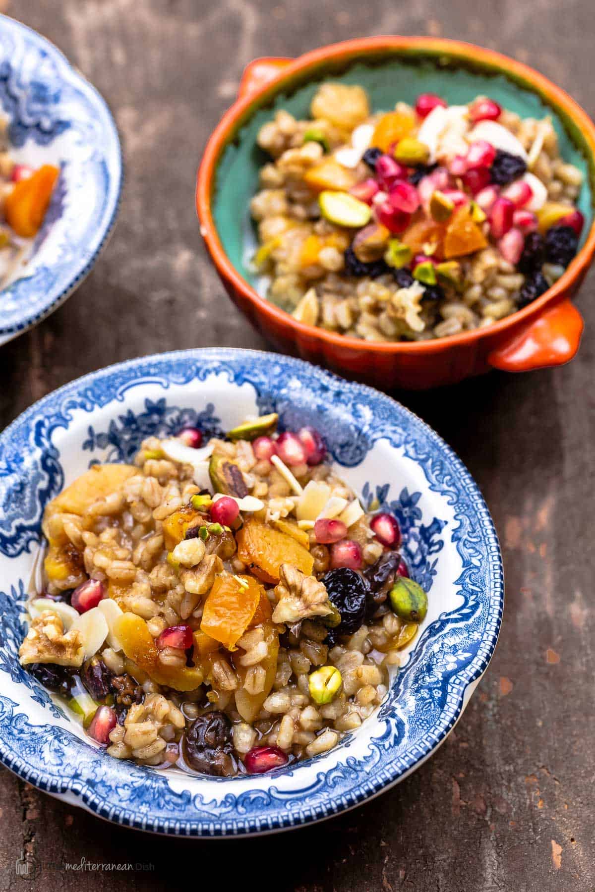 burbara (wheat berry pudding) in a blue and white bowl topped with pomegranate seeds and nuts. Additional servings in the background