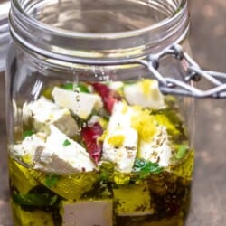 side view of marinated feta in a jar with olive oil, dry chiles, jalapeno, orange zest, and herbs