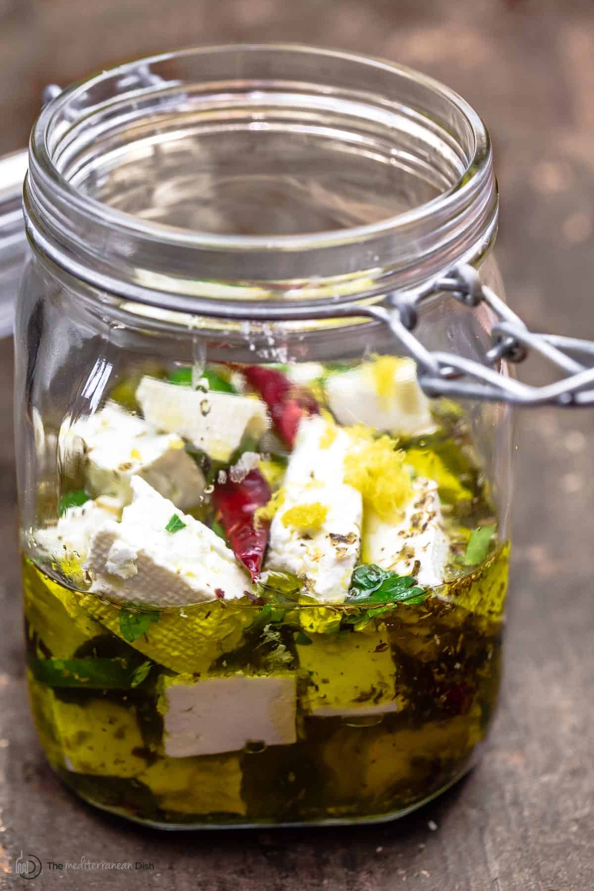 side view of marinated feta in a jar with olive oil, dry chiles, jalapeno, orange zest, and herbs