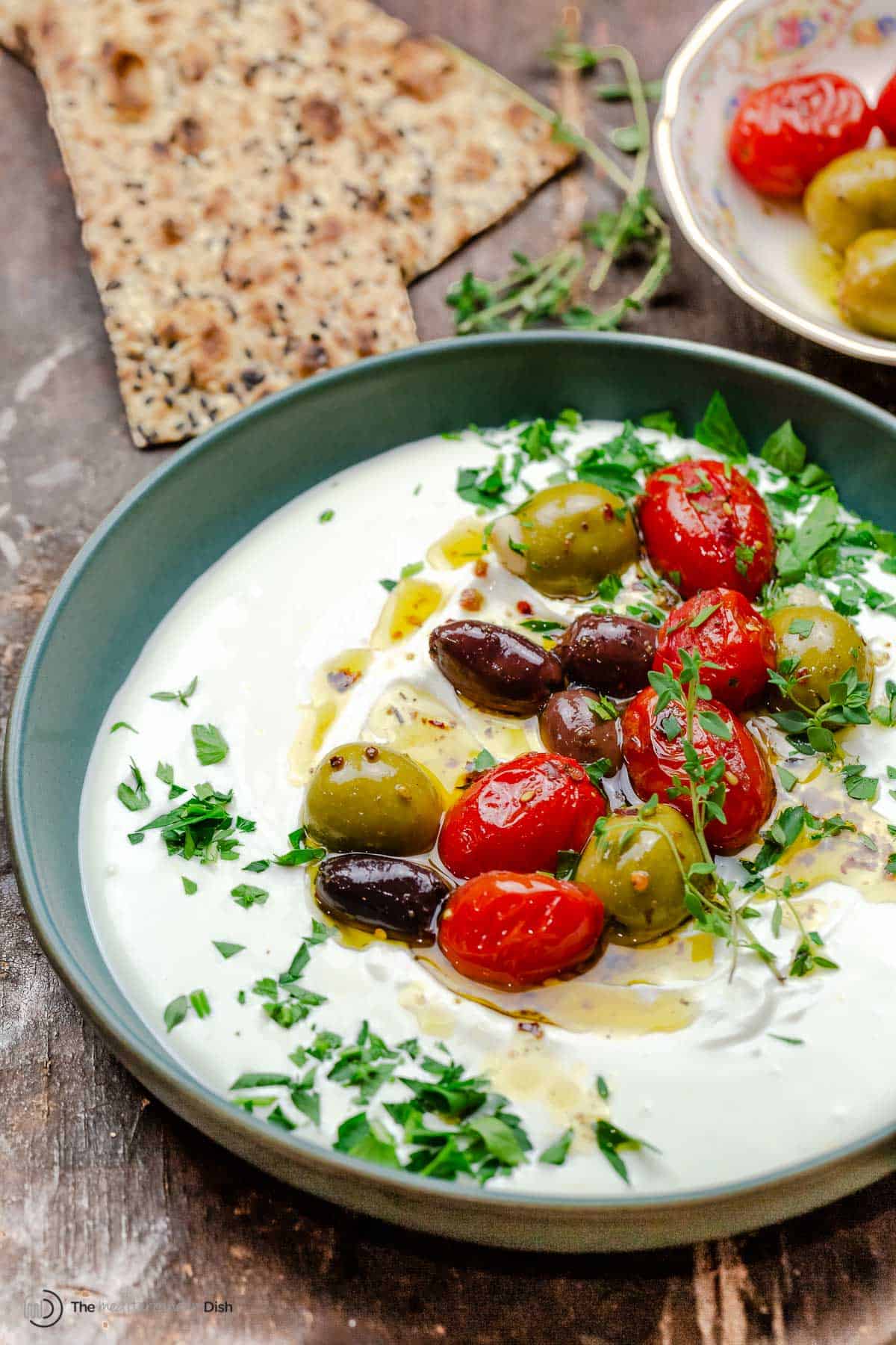 whipped labneh dip in a bowl topped with warmed olives and tomatoes in olive oil, garnished with parsley. Crackers in the background