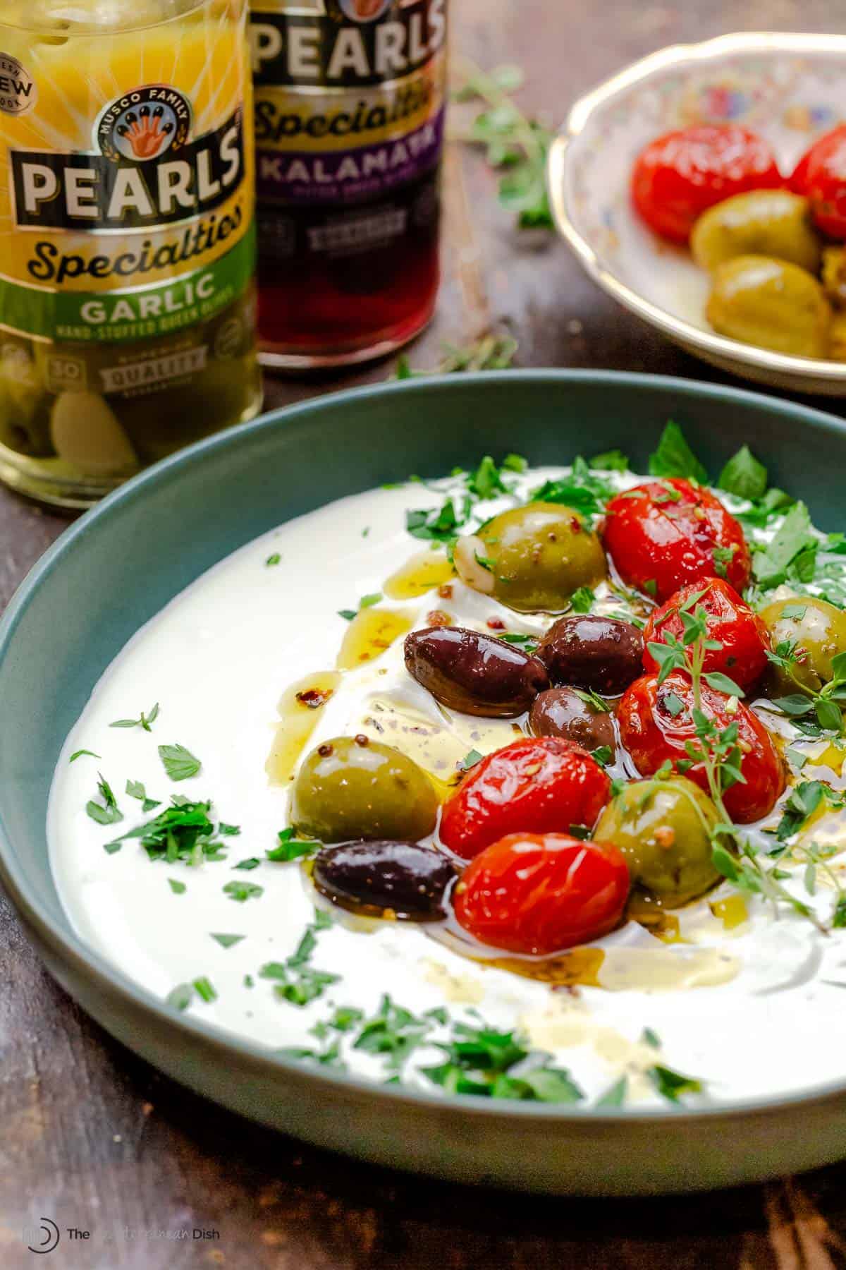 whipped labneh dip in a bowl topped with warmed olives and tomatoes in olive oil, garnished with parsley. situated next to two jars of pearls olives