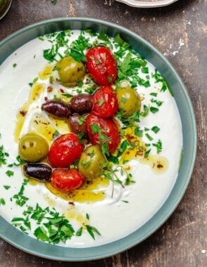 whipped labneh dip in a blue bowl topped with warmed olives and tomatoes in olive oil, garnished with parsley