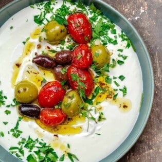 whipped labneh dip in a blue bowl topped with warmed olives and tomatoes in olive oil, garnished with parsley