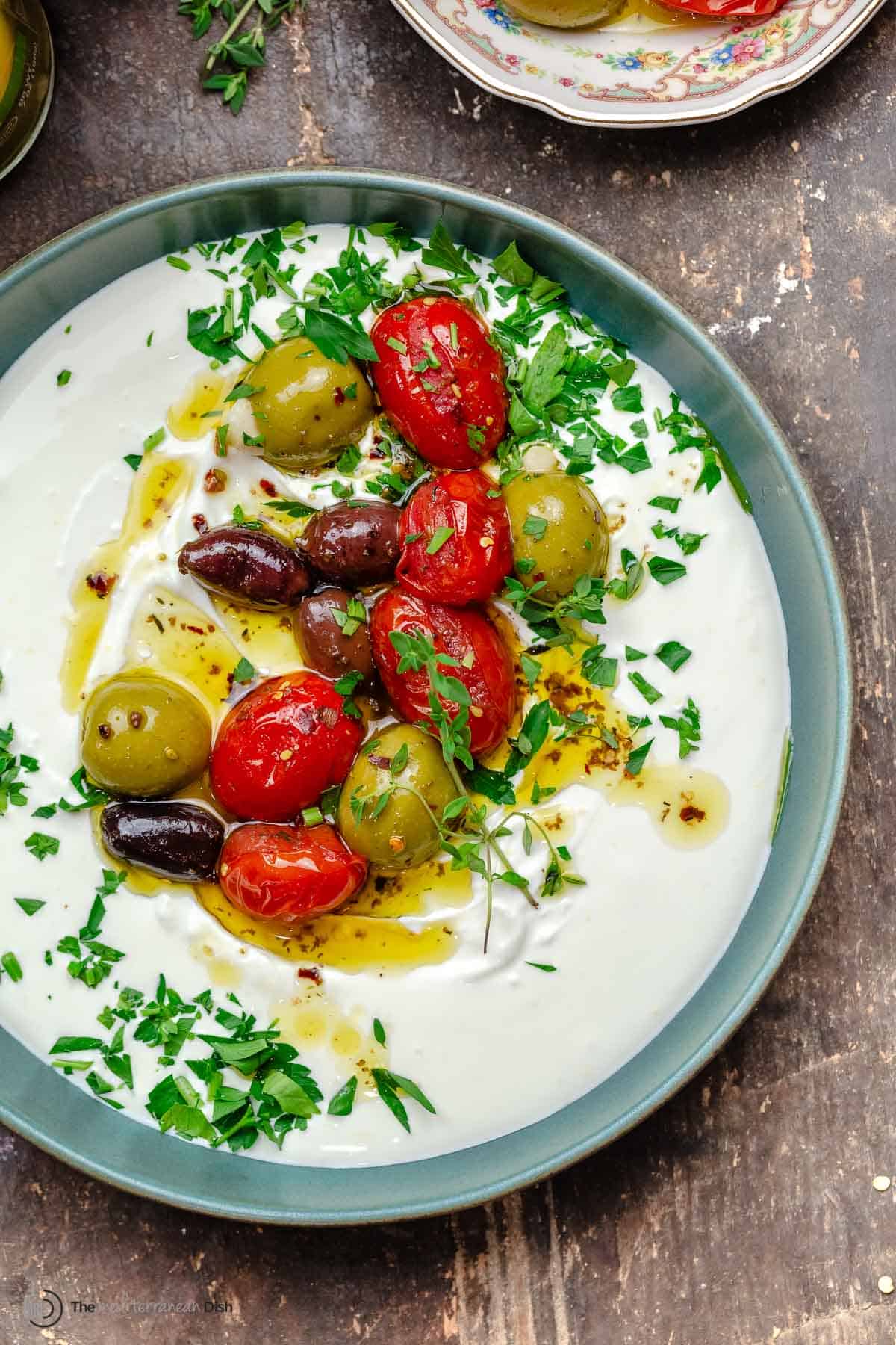 whipped labneh dip in a blue bowl topped with warmed olives and tomatoes in olive oil, garnished with parsley