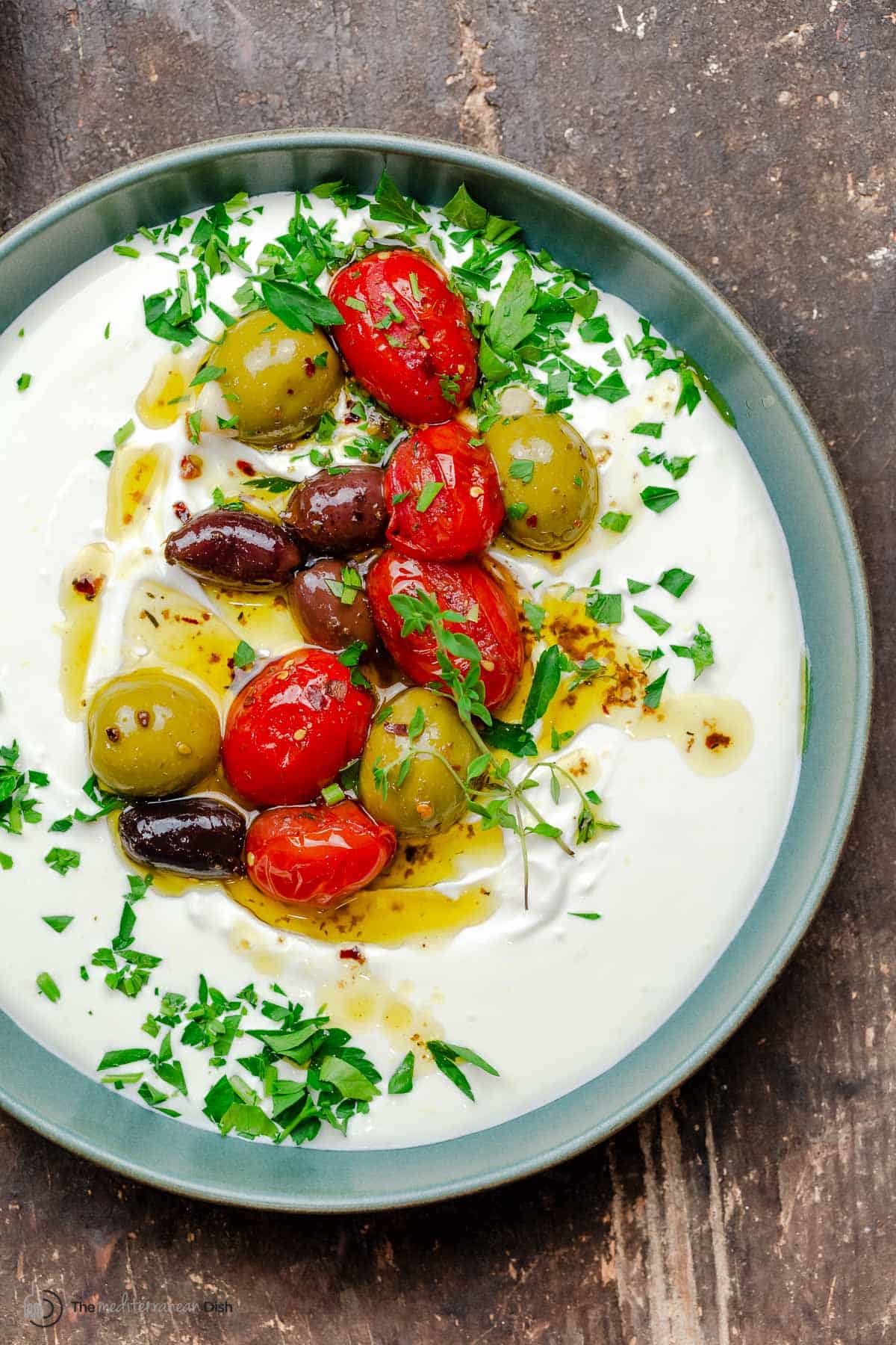 whipped labneh dip in a bowl topped with warmed olives and tomatoes in olive oil, garnished with parsley