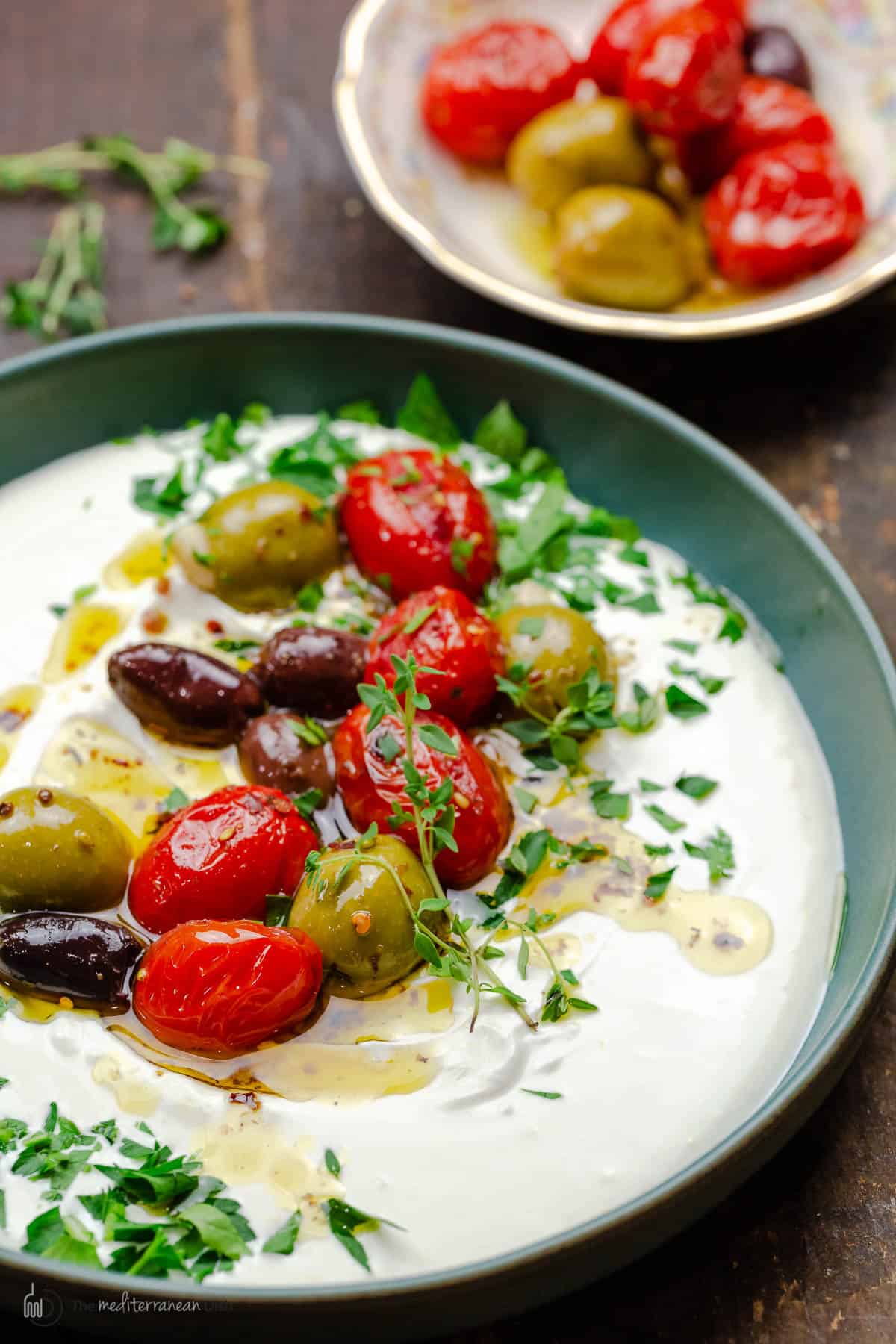 whipped labneh dip in a bowl topped with warmed olives and tomatoes in olive oil, garnished with parsley. another bowl of warmed olives and tomatoes in the background