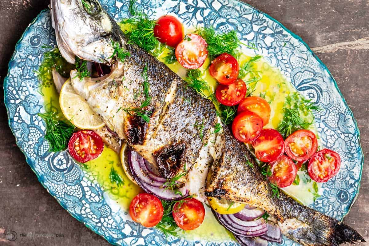 broiled branzino on a platter, bathed in ladolemono, with halved cherry tomatoes and dill sprigs on the side