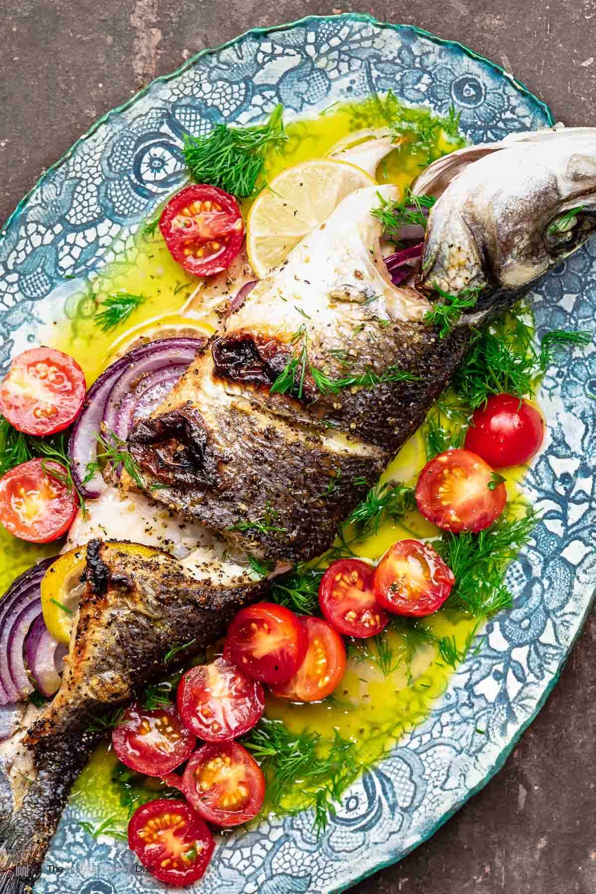 branzino stuffed with red onion and lemon on a platter with ladolemono, cherry tomato halves, and dill
