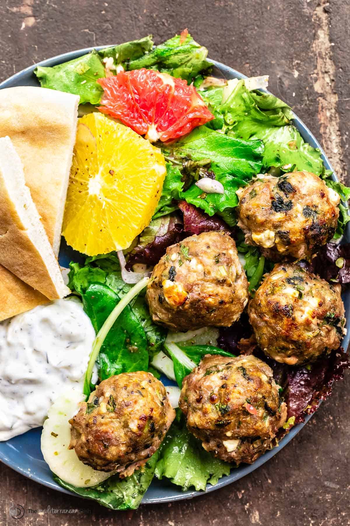 Baked turkey meatballs served on a blue plate over a salad with a side of tzatziki sauce and pita bread