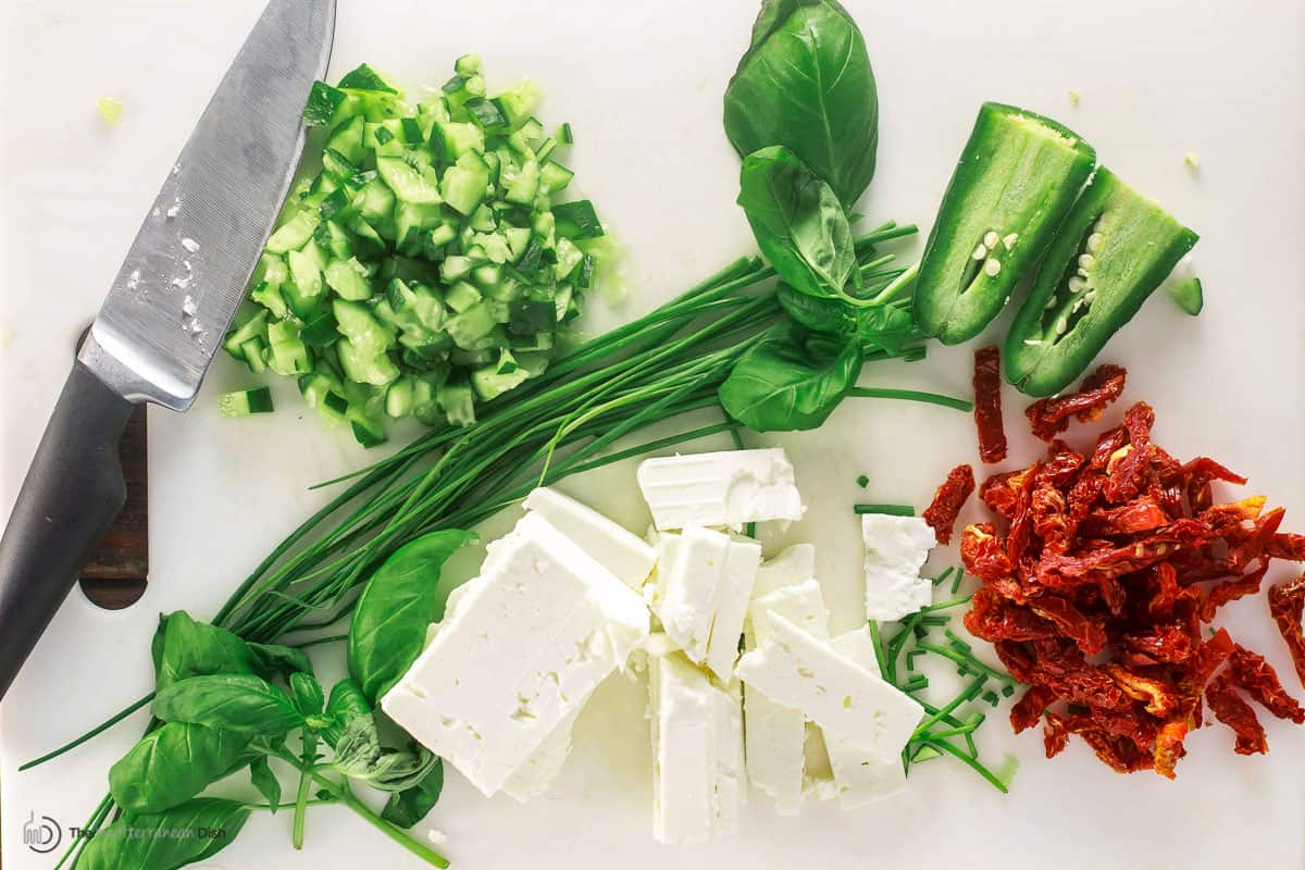 ingredients for feta dip including feta cheese, chopped cucumber, basil leaves, chives, jalapeño, and sun-dried tomato bits