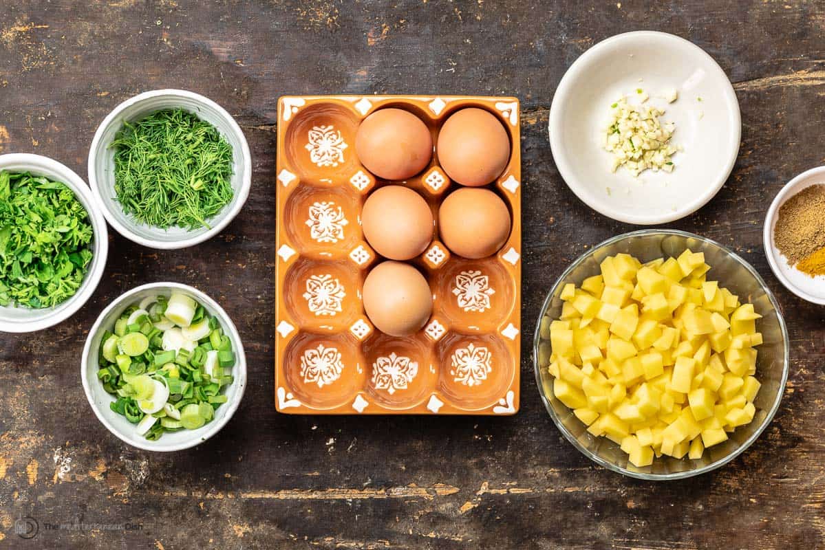 ingredients for potato omelet including eggs, gold potatoes, spices, minced garlic, fresh parsley and dill, and green onions