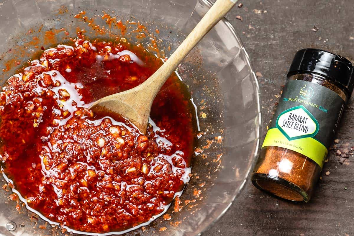 baharat chicken marinade in a glass bowl with a wooden spoon. The Mediterranean Dish baharat spice blend is next to the bowl.