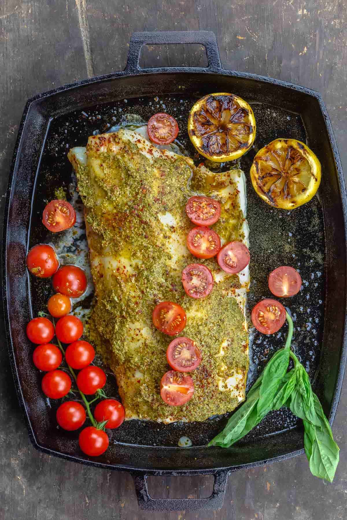 baked Chilean sea bass with basil pesto in a large cast-iron baking dish. Topped with cherry tomatoes, with a sprig of fresh basil on the side